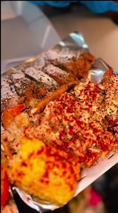 Foil tray holding seasoned wings, shrimp, corn and a loaded seafood-topped portion